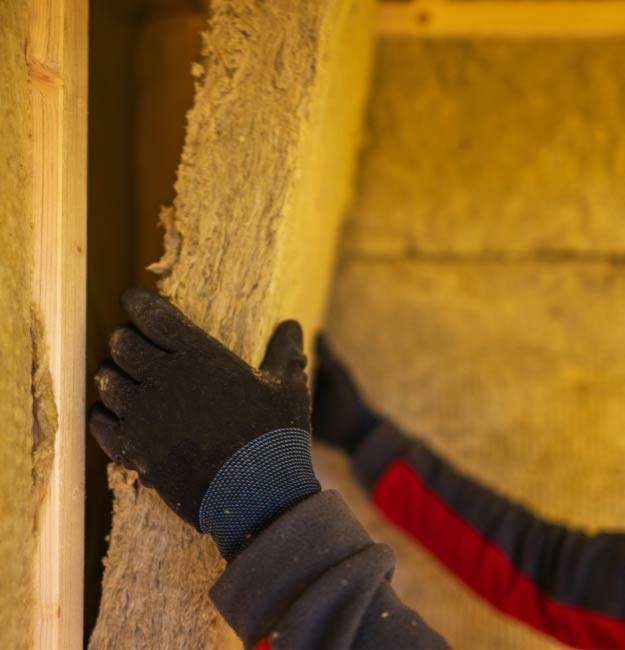 Close-up of a worker wearing black gloves and a red-sleeved jacket fitting a dense, fibrous mineral wool insulation panel into a narrow wall cavity.