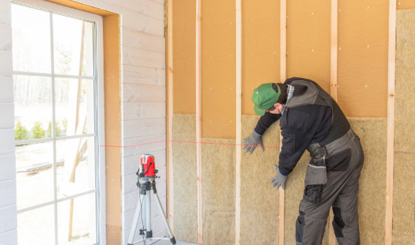 A worker in a green cap and black workwear presses mineral wool insulation into a wooden stud wall inside a partially constructed room, with a window and laser level visible nearby.