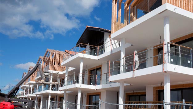 A multi-story apartment complex under construction, with exposed wooden framing on the upper floors and white concrete lower floors, scaffolding, and workers visible.