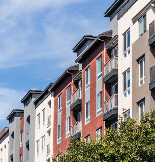 A modern multifamily apartment building with a mix of red brick and gray stucco exterior, featuring large windows and private balconies against a clear blue sky.
