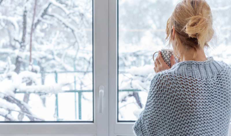 Woman holding a hot drink while looking out a window at a snowy winter landscape.